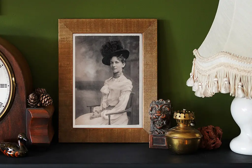 A framed black-and-white portrait of a woman in Edwardian clothing and a large hat sits on a shelf, surrounded by a clock, a vintage lamp with a fringe, small figurines, and pinecones against a dark green wall.
