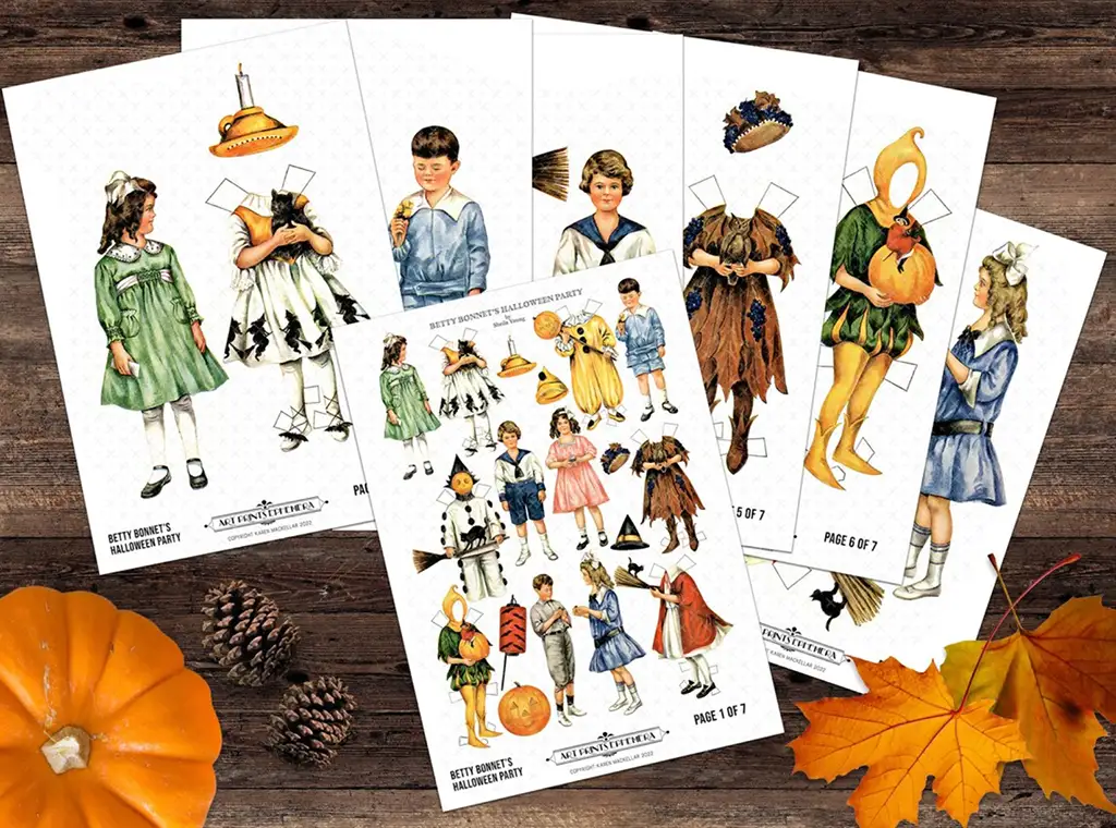 A flat lay of six sheets featuring vintage-style cut-out paper dolls in various costumes, including animals, fairy tale characters, and Halloween-themed outfits. A small pumpkin, pine cones, and autumn leaves adorn the wooden background.