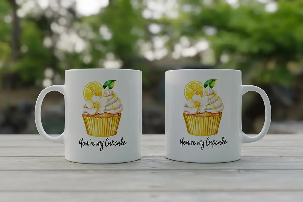 Two white mugs sit on a wooden table outdoors, each featuring an illustration of a lemon cupcake topped with cream and garnished with a lemon slice. The text on the mugs reads "You're my Cupcake." The background is blurred greenery.
