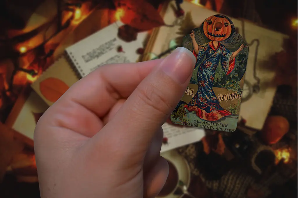 A close-up of a hand holding a vintage Halloween card with a pumpkin-headed figure in a colorful robe. The background is blurred but shows an autumn scene with leaves, warm lights, and what appears to be an open book with handwritten text.