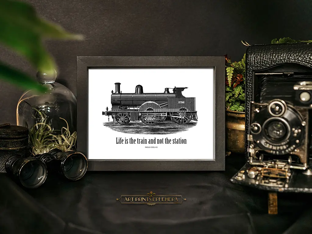Framed black-and-white image of a vintage steam train with the text "Life is the train and not the station" on a dark table. Surrounding items include an old camera, binoculars, a potted plant, and a protective glass dome containing air plants.