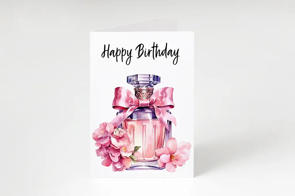 A birthday card features an elegant perfume bottle adorned with a pink ribbon and surrounded by pink flowers. The text "Happy Birthday" is written in a stylish font at the top of the card. The background is plain white.