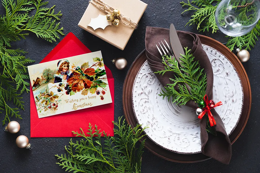 A festive setting includes a holiday greeting card in a red envelope, a wrapped gift, greenery, and a place setting. The plate is decorated with a brown napkin, silverware, and a sprig of greenery, with a wine glass and Christmas ornaments nearby.