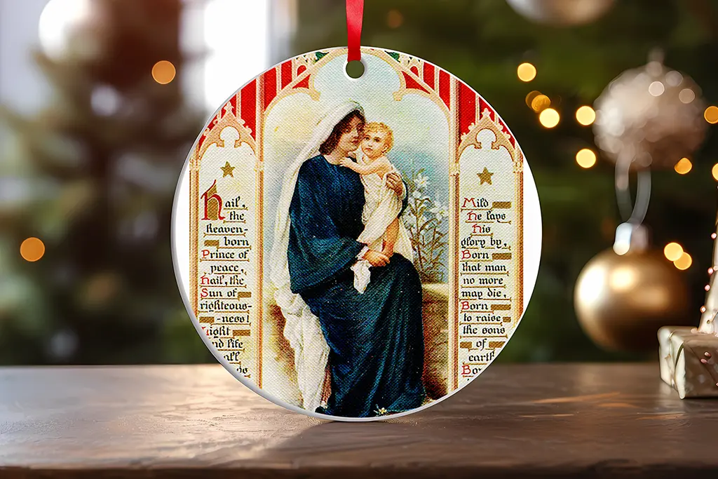 A round ornament featuring a classical painting of the Virgin Mary holding baby Jesus, adorned with a red ribbon for hanging. The background includes text with religious phrases and Christmas decorations, including a blurred Christmas tree and baubles.