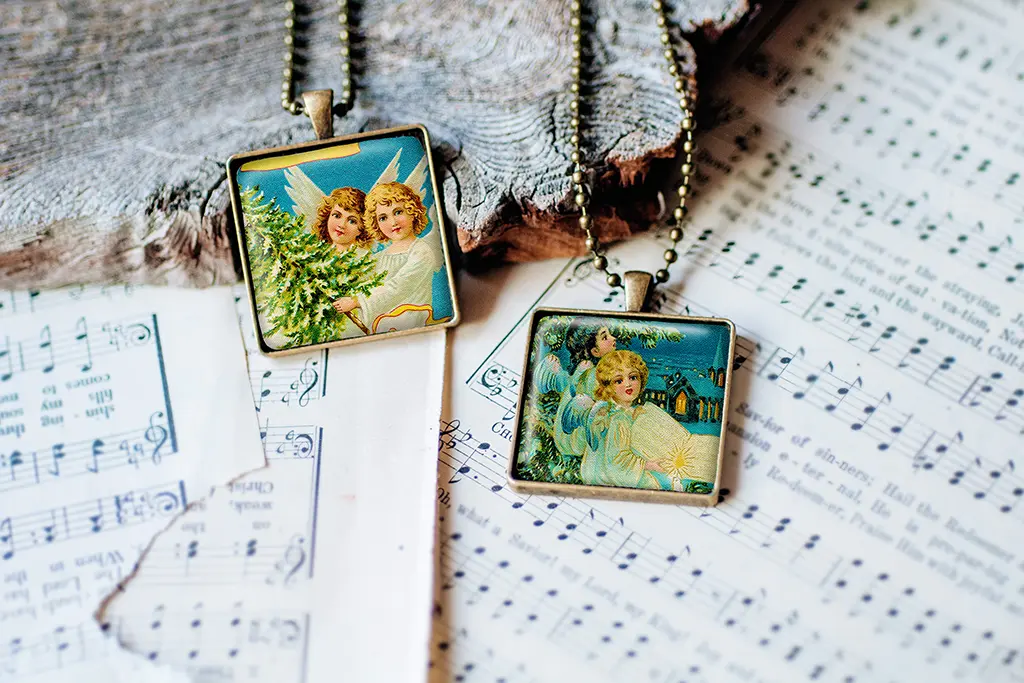 Two square pendants with vintage-style angel illustrations rest on sheet music. One pendant shows two angels beside a Christmas tree, and the other depicts an angel in a blue robe against a wintry scene. Both hang from metallic ball chain necklaces.