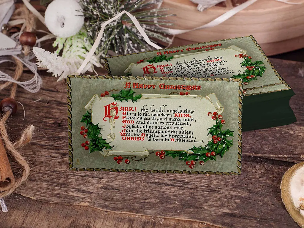A Christmas card on a wooden surface with festive decorations in the background. The card features the title "A Happy Christmas" and decorative holly illustrations. There is Christmas-themed text in a medieval-style font. The background includes pinecones and candles.